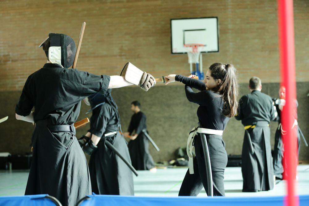 A black belt with a fencing mask on the left is correcting the posture of a female white belt on the right.